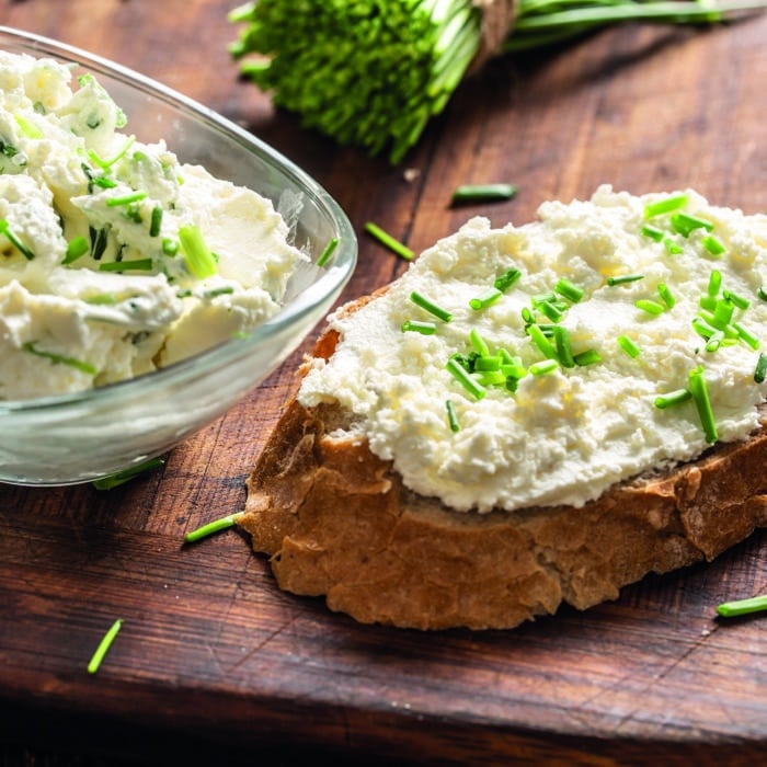 A bowl of homemade cream cheese spread with chopped chives surrounded by bread slices with spread and a bunch of freshly cut chives